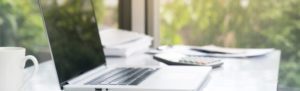 Photograph of a laptop, a coffee mug, a calculator, and a stack of papers on a desk, with a view of greenery outside the window in the background.