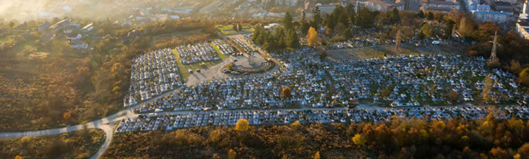 Aerial view of a large cemetery situated in an urban area, surrounded by trees, roads, and buildings. The cemetery is densely packed with graves, with a circular monument visible near the center. The landscape transitions from the cemetery into surrounding greenery and urban development.