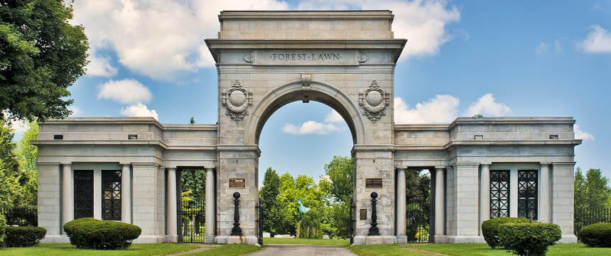 Photograph of the grand entrance archway at Forest Lawn Cemetery, featuring a large stone arch with the name 'Forest Lawn' engraved at the top. The structure is flanked by columns and ornate gates, with lush green trees and a blue sky visible in the background.