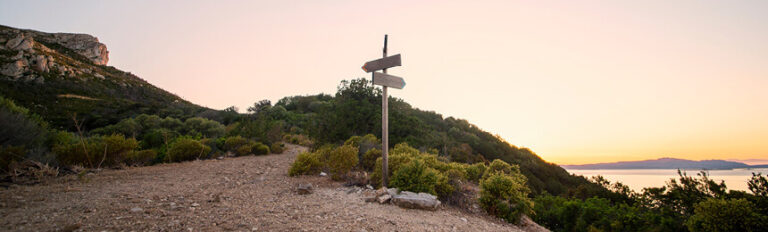 Landscape at sunset showing a dirt path leading to a wooden signpost in the middle of a natural, bushy area with hills in the background. The sky is painted in soft pastel colors as the sun sets over the distant water horizon.