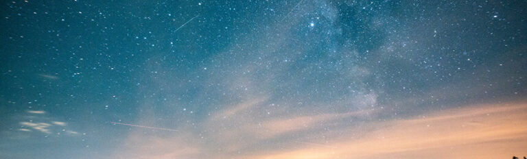 Night sky filled with stars and streaks of light from distant shooting stars, with soft clouds illuminated by the last light of sunset near the horizon.