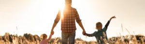 Photograph of a person walking hand in hand with two children through a sunlit field at sunset. The figures are silhouetted by the golden light, with tall grasses surrounding them, creating a warm, nostalgic atmosphere.