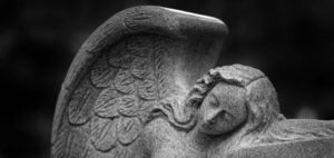 Close-up photograph of an angel sculpture in a cemetery, carved from stone, with the angel resting its head on folded arms and detailed wings visible in the background. The image is in black and white, adding to the somber tone.